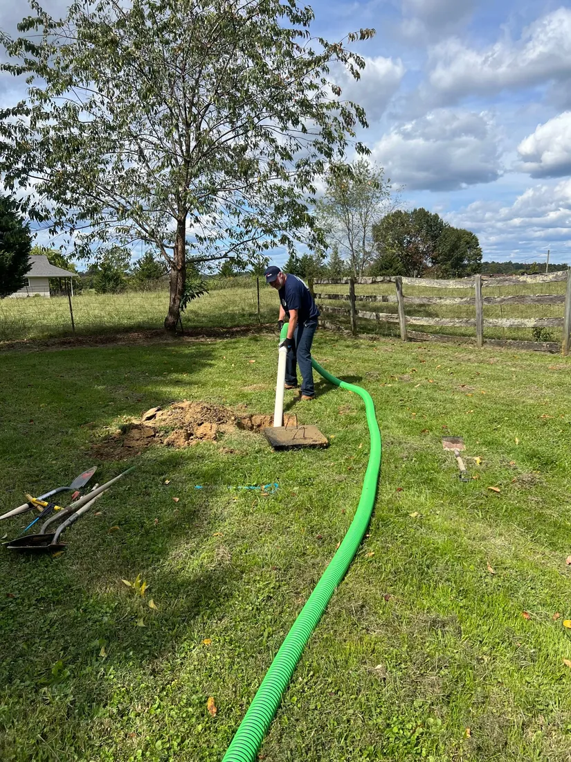 A Grace Septic technician works at an uncovered tank lid in a fenced pasture, with the hose running back across the grass.