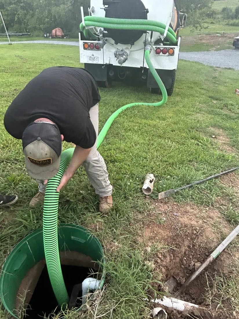 A technician feeds the green suction hose into an open septic tank riser while the truck waits in the field behind him.