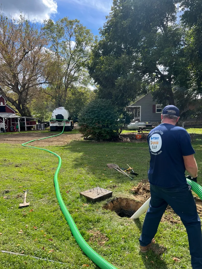 A Grace Septic technician in a navy company shirt holds the hose at an open septic tank in a Hawkins County yard, with the truck parked by the trees.